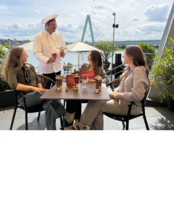 schokoladenmuseum-koeln-grand-cafe-aussen Drei Frauen werden im Chocolate Grand Café im Schokoladenmuseum Köln auf der Außenterasse von einem Chocolatier bedient.