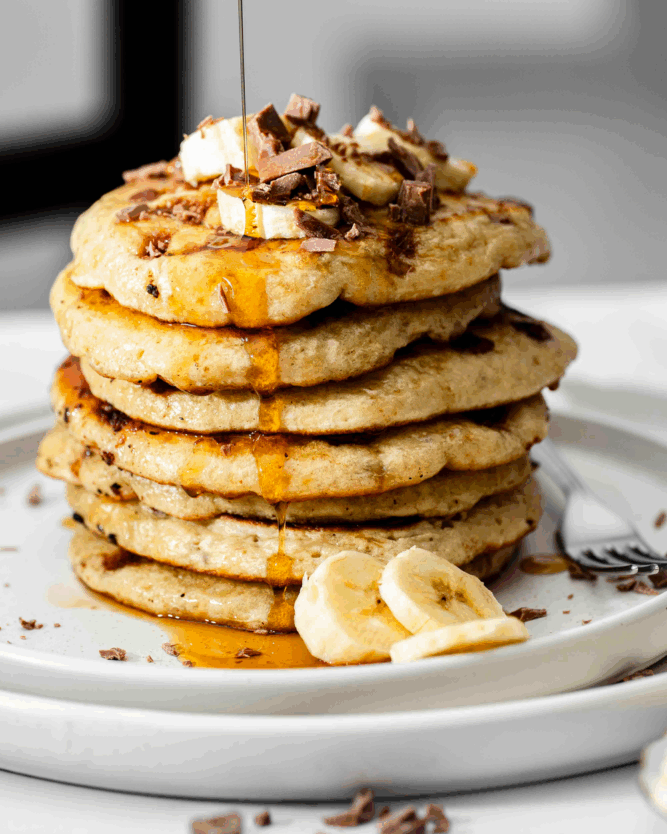 Als Turm gestapelte Chocolate Chip Banana Pancakes von VIVANI, die mit Ahornsirup beträufelt werden.