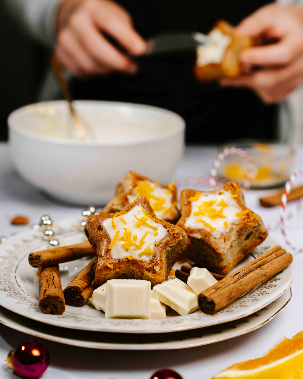 Zimtsternküchlein mit weißer Schokoladenglasur auf einem Teller mit Zimtstangen und weißen Schokoladenstückchen.