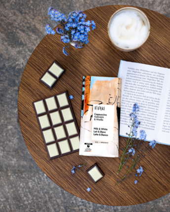 Cappuccino-Schokolade von VIVANI mit aufgeschlagenem Buch, einer Tasse Cappuccino und Blumen auf einem Tisch.