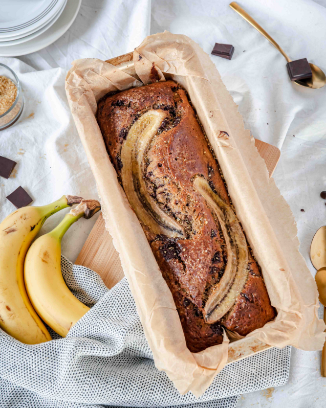 Bananenbrot in einer Kastenform mit einem Strunk Bananen und Schokostückchen auf einer Tischdecke.