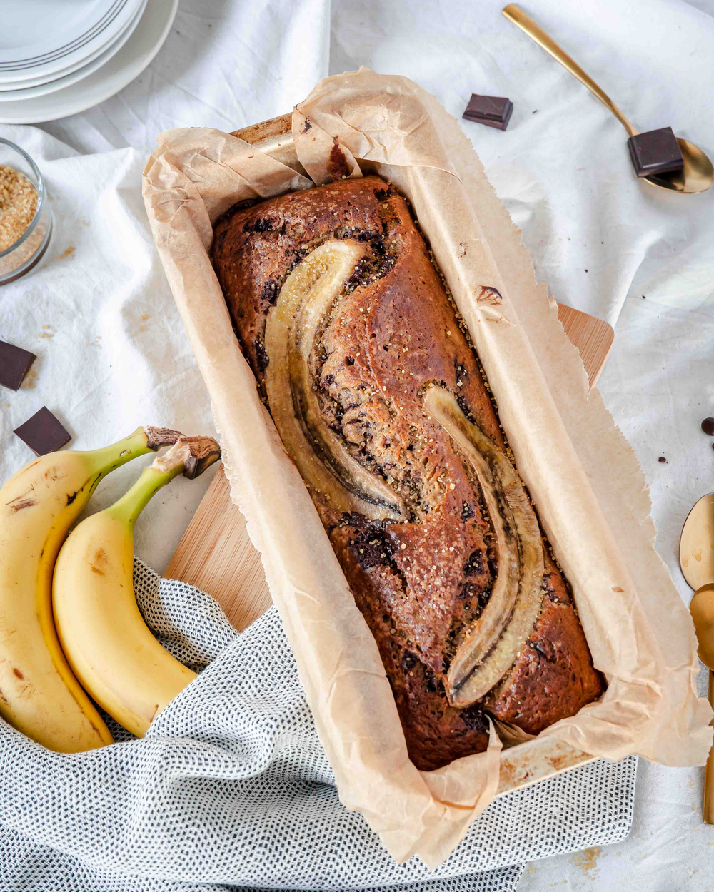 Bananenbrot in einer Kastenform mit einem Strunk Bananen und Schokostückchen auf einer Tischdecke.