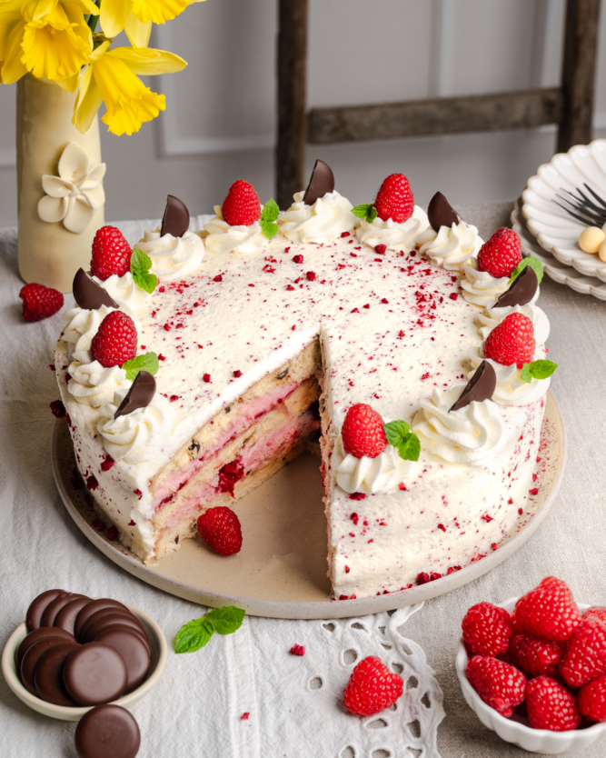 Eine angeschnittene Sahnetorte mit mehreren Schichten, Himbeeren und Schokoladenblättchen auf einem Tisch angerichtet.