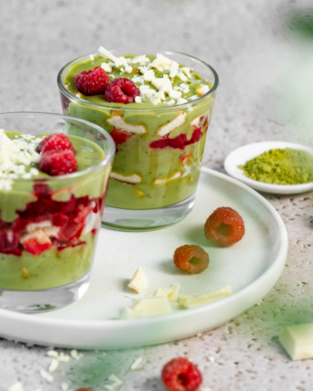 matcha-himbeer-tiramisu Zwei Dessertgläser mit Matcha-Himbeer-Tiramisu und Himbeeren, Matcha-Pulver und weißen Schokoladensplittern als Dekoration.