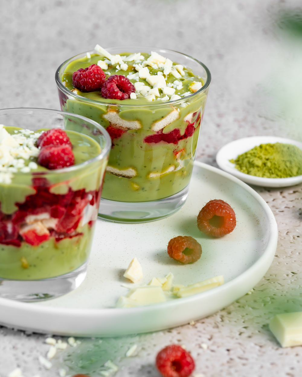 Zwei Dessertgläser mit Matcha-Himbeer-Tiramisu und Himbeeren, Matcha-Pulver und weißen Schokoladensplittern als Dekoration.