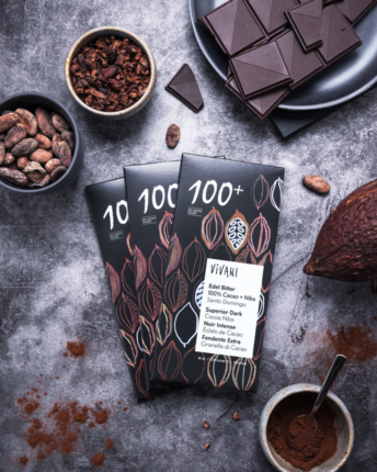 edel-bitter-100-prozent-cacao-plus-nibs-vivani-bio-schokolade Drei Tafeln Edel Bitter 100 % Cacao + Nibs von VIVANI Bio-Schokolade, mit Kakaobohnen und anderen Zutaten auf einem Tisch dekoriert.